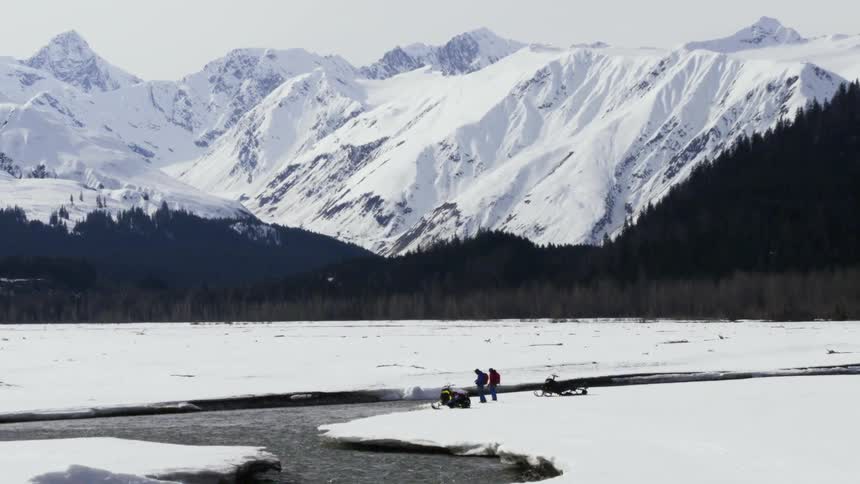 Aerial of two people next to snowmobiles by a mountain river
