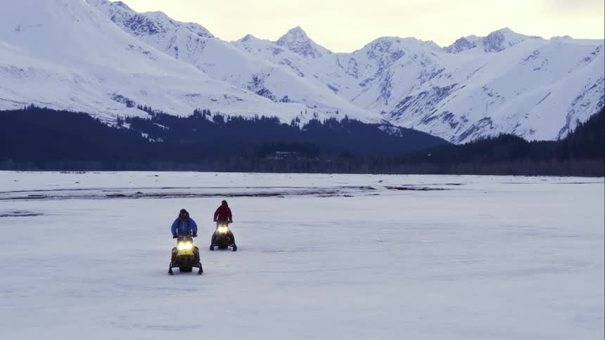 Aerial of two people on snowmobiles riding across frozen lake in front of mountains