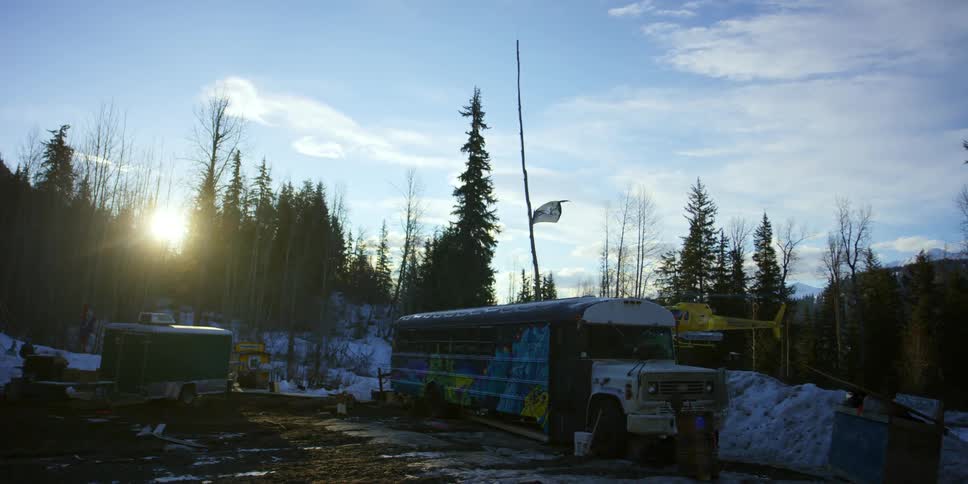 Sun peaks over forest highlighting parked bus as helicopter rises into air