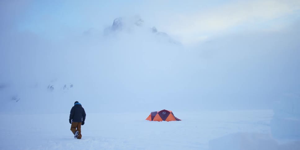 A man walks toward and around an orange tent set up near mountains in the arctic