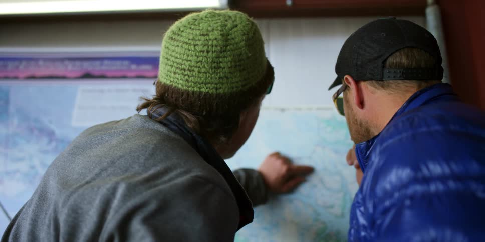 Two men with hats and sunglasses talking and pointing to a map on the wall