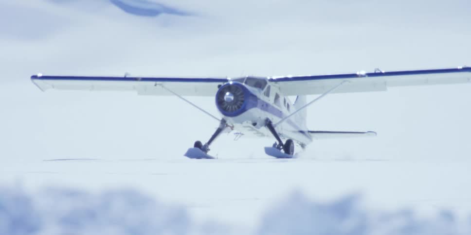A pilot lands a small plane on snow ground between mountains and near a tent