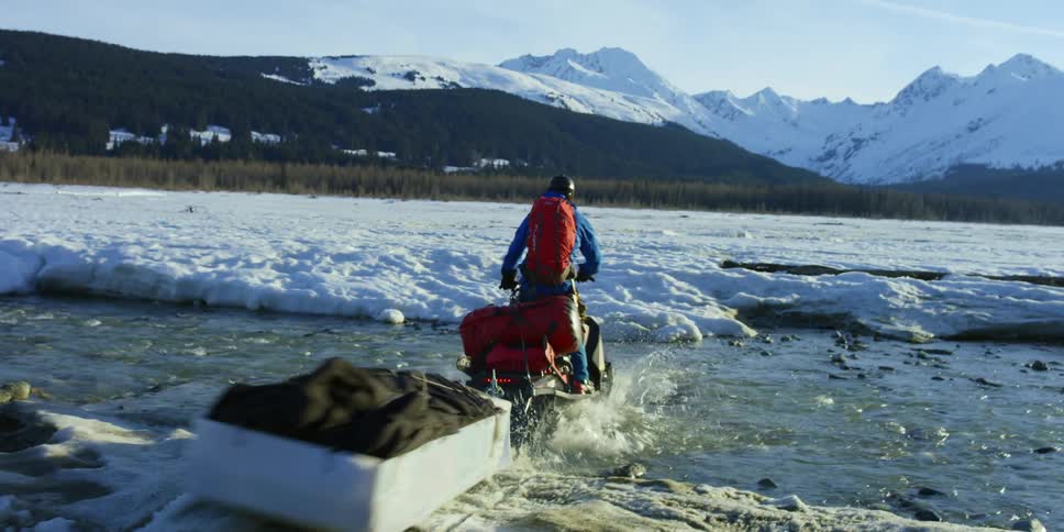 A person rides an all terrain vehicle over ice covered terrain and then into a creek