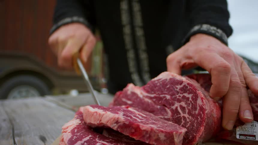 A man uses a large knife to cut through steak on an outside wood table