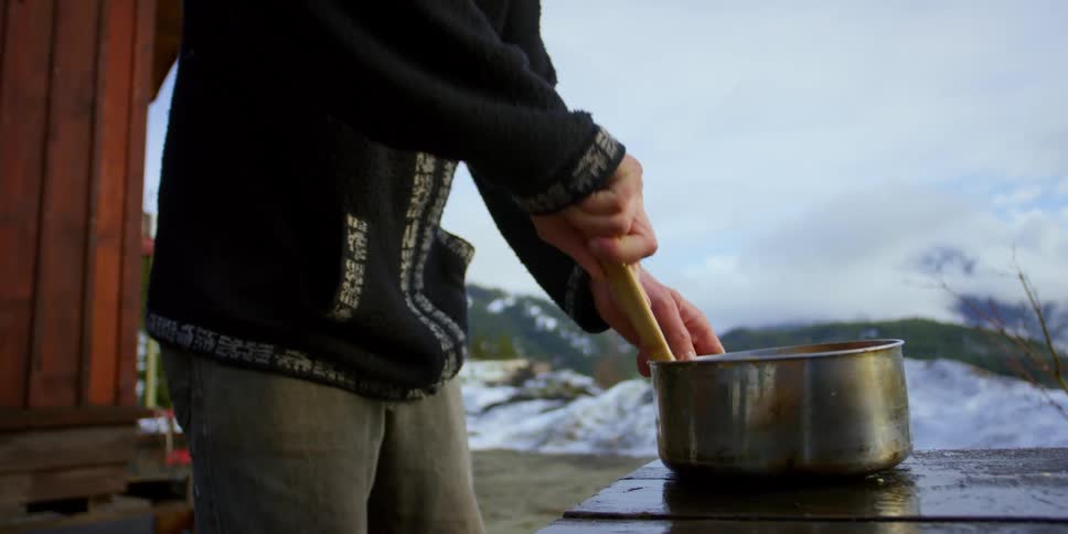 Man stirs cooking pot on picnic table outside cabin