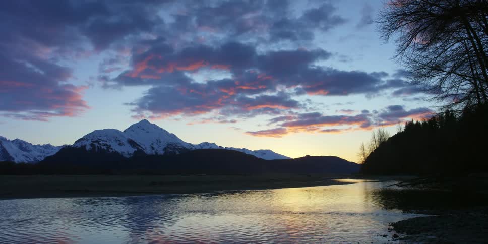 A gently flowing river through a field at the base of a snow capped mountain in the evening
