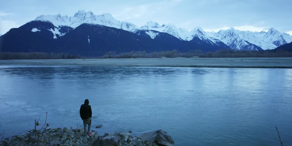 Man stands on the shore of a river near the snow covered peaks of a mountain range at morning