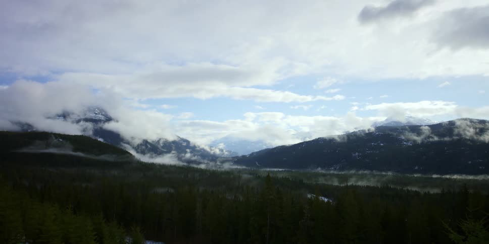 Clouds pass over an evergreen forest at the base of mountain ridge