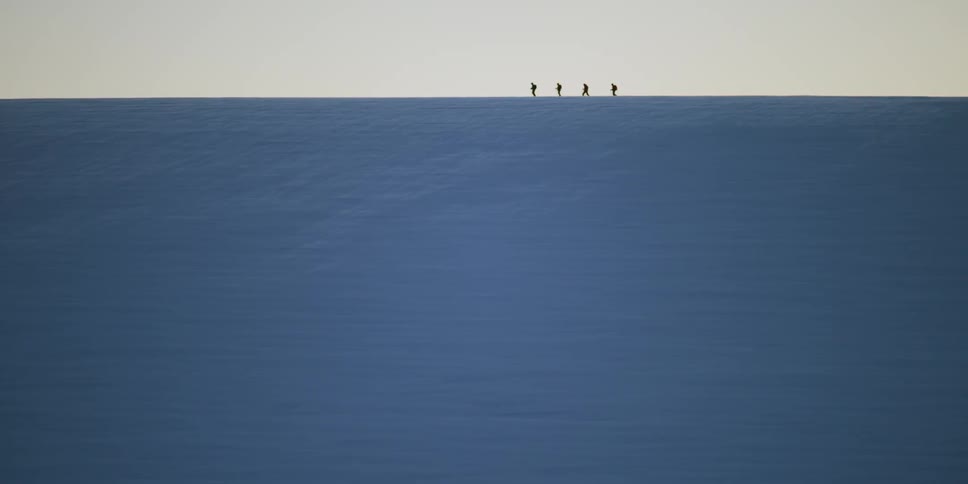 A silhouette of four people hiking on a snowy plane with the sky and clouds in the background