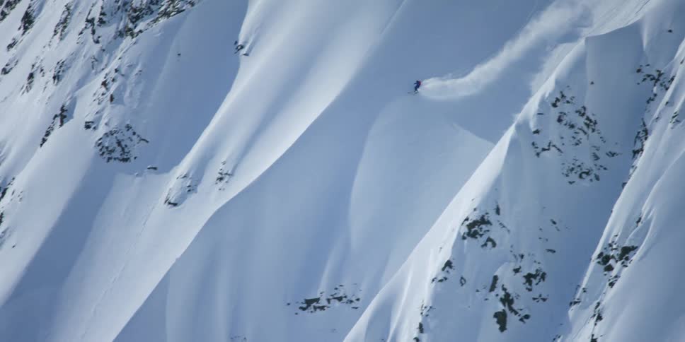 A skier descends a steep mountain slope covered in deep snow on a sunny day