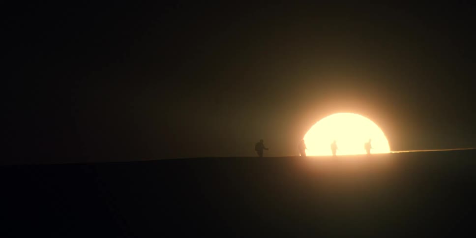 The silhouettes of hikers cross the top of a hill as the low sun in a black sky peaks over the horizon