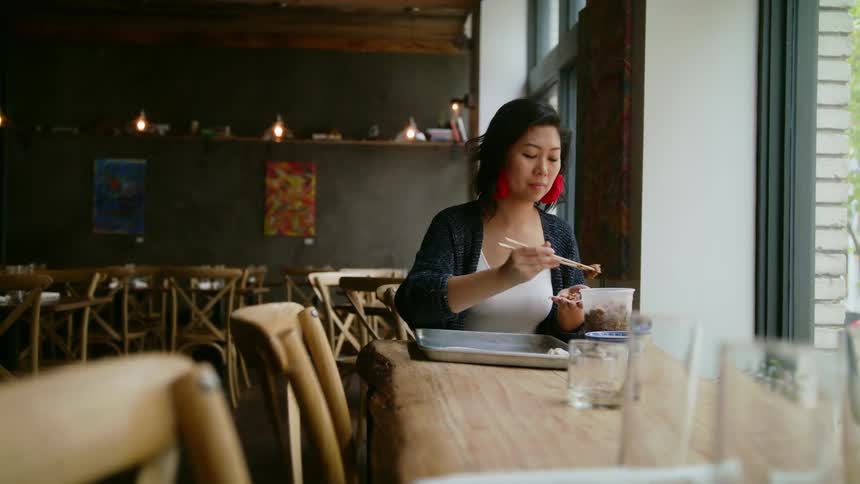 A woman uses chopsticks to pick up food sitting in a restaurant by herself