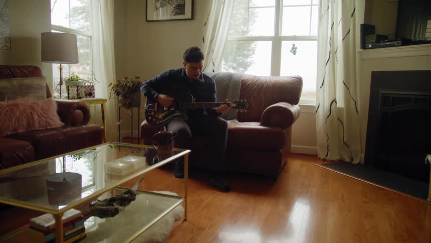Man with a goatee beard sits on a brown couch and strums a guitar in the living room