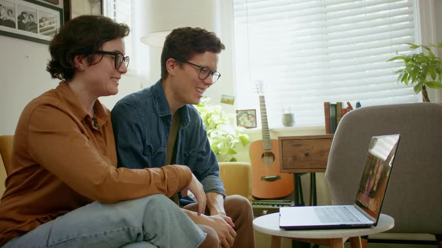 A man and woman sit on a couch and wave to a laptop having a video call