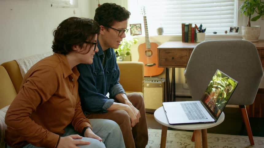 A young man and woman at home wave and video chat with a couple with their baby