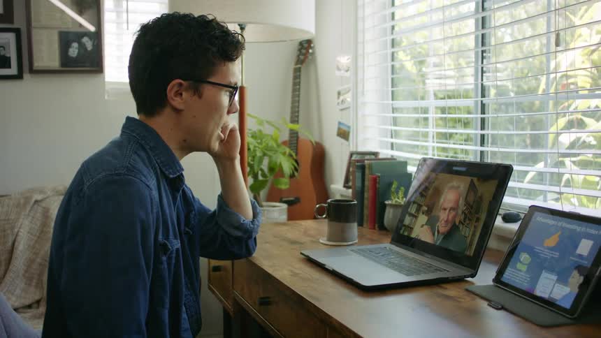 A young man sits at a desk at home and talks to an older man via video call