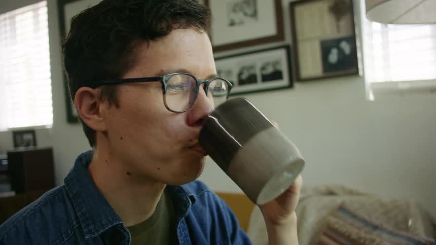 Young man wearing glasses drinks a hot beverage from a cup in a room at home