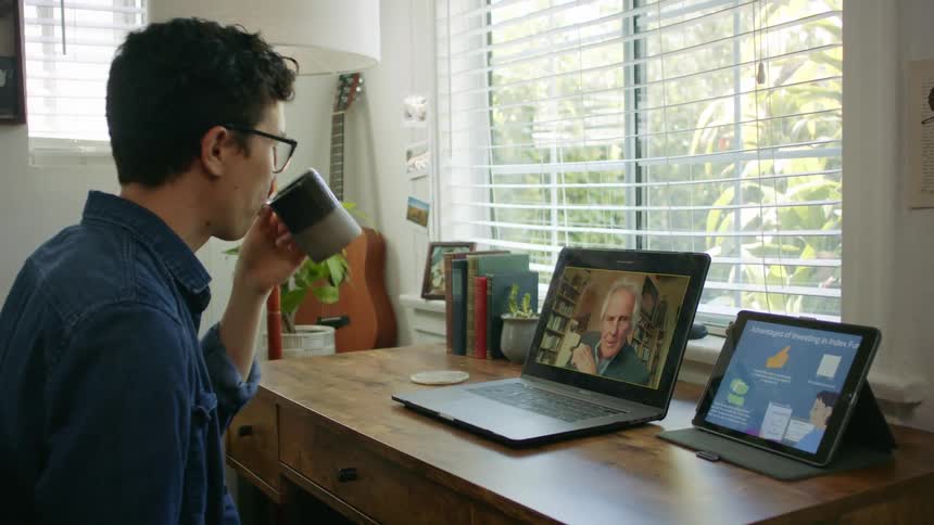 Male sitting at desk drinks a cup of coffee in front of a computer and tablet while video chatting with elderly male on computer screen