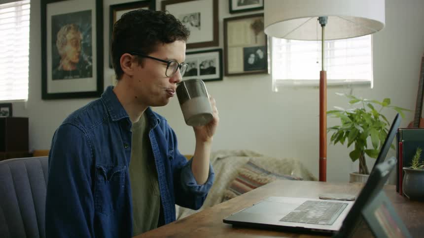 A man wearing glasses sits at his desk and smiles as he chats with someone via his laptop