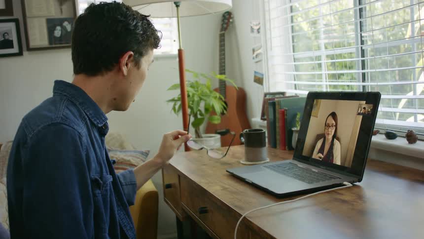 A man takes off his eye glasses and rubs his eyes as he particpates in a video conference with a woman on his laptop