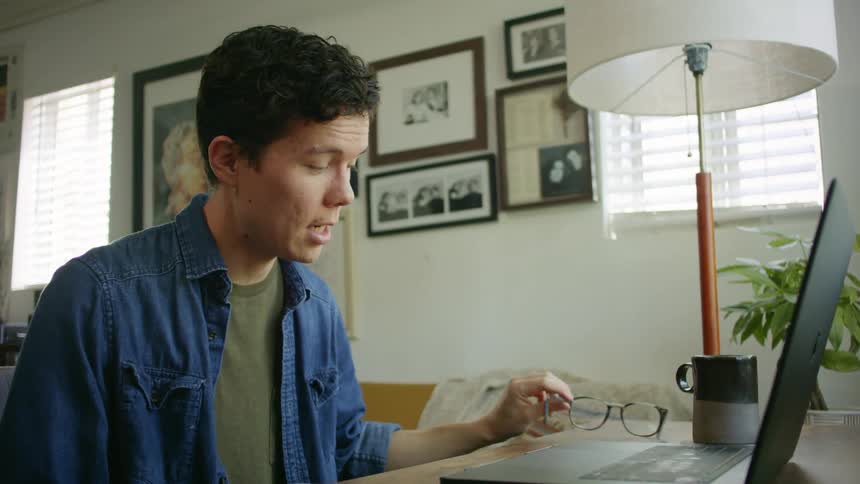A young man removes his glasses as he sits at a desk speaking into the microphone of his laptop