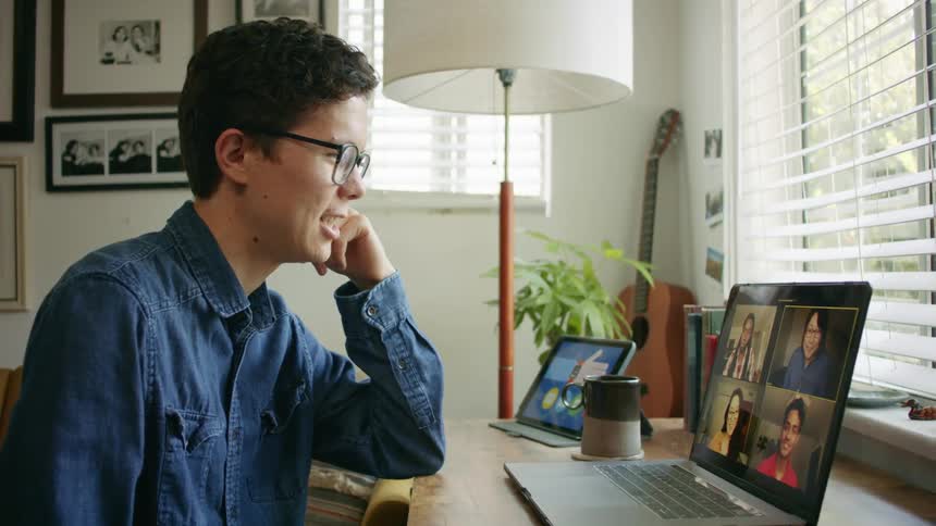 A man sits at a small desk having a video call on a laptop