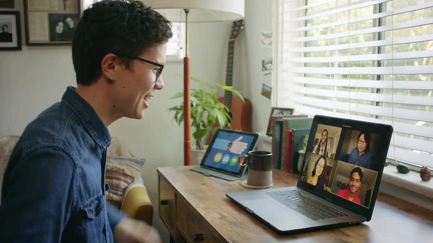 A man sits at a small table having a video call with multiple people
