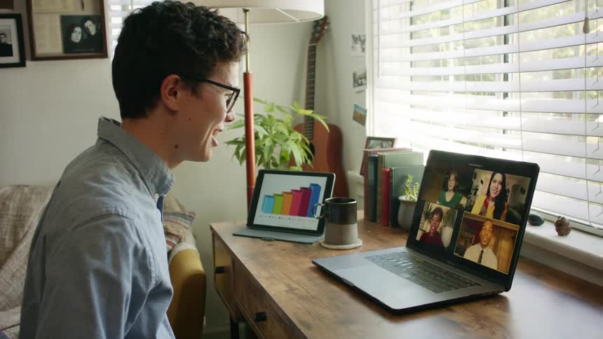 A man sits at a small desk in front of a laptop having a video call