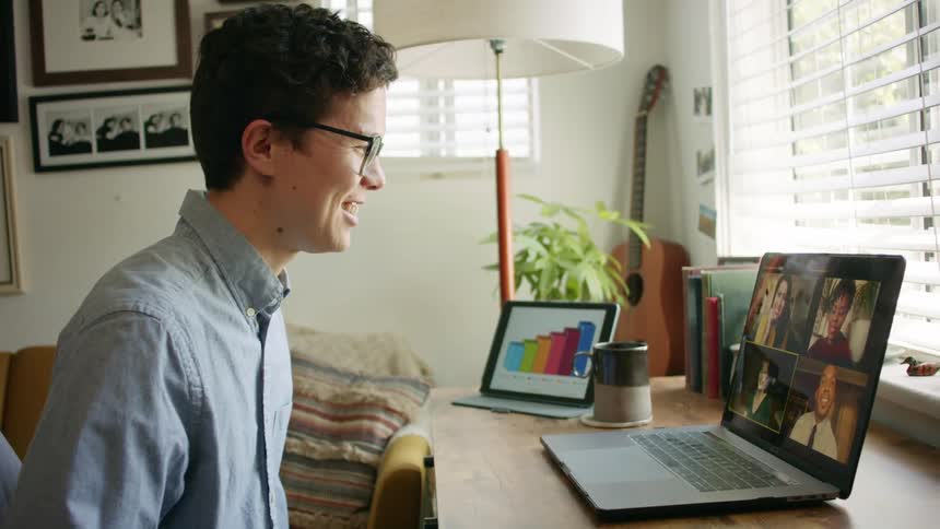 A man sits at a small desk with a laptop having a video call