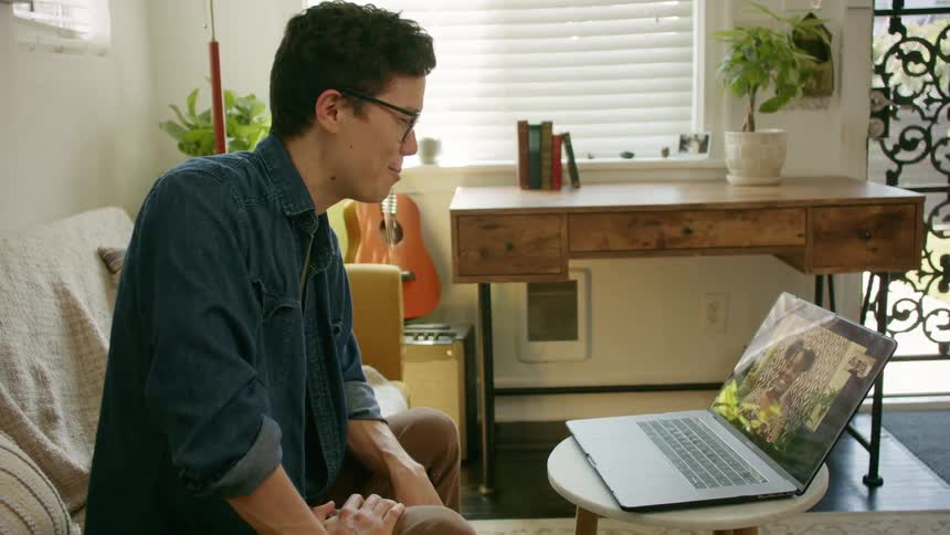 Man with glasses and a bluet shirt sits and makes a video call on macbook pro with black woman in the living room at home