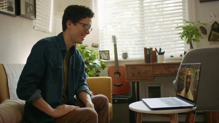 A young man sits on a living room sofa while video chatting on a laptop