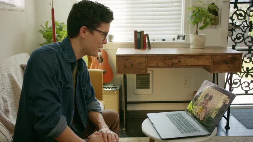 A young man at home waves as he video chats with a woman on a laptop