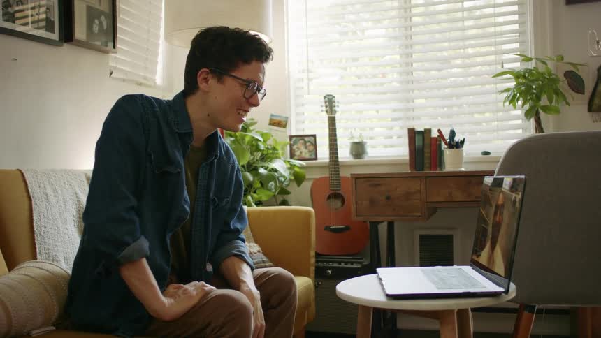 Male sitting on sofa in front of laptop resting on a round table laughs and waves while video chatting to male on computer screen