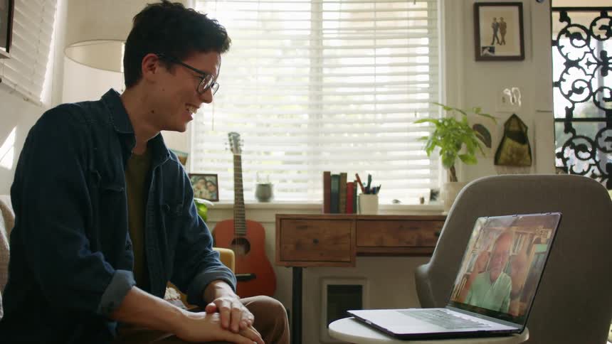 Side view of a man with glasses and a blue shirt making a video call with an elderly man in the living room at home