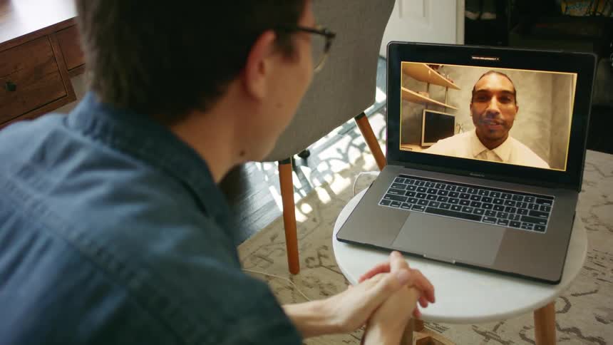 A man waves at his friend and laughs as they talk in a video call on his laptop