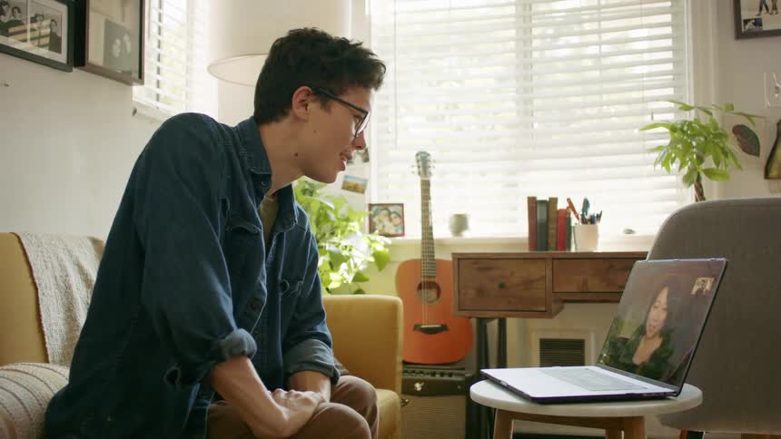 A man sitting in a couch in front of a laptop on a stool video chatting