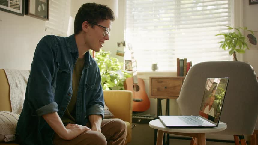 A young man sits on his couch at home as he waves at his laptop while video chatting with someone