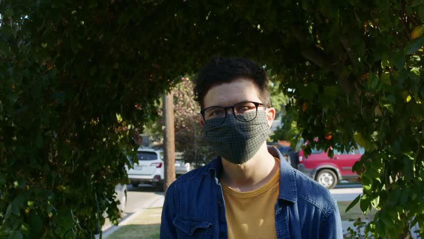 A young man in a medical mask looks into the camera through a hole in a hedge