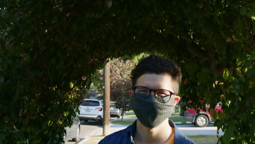 A young man in a mask blinks as he looks into the camera through a large hole in a hedge