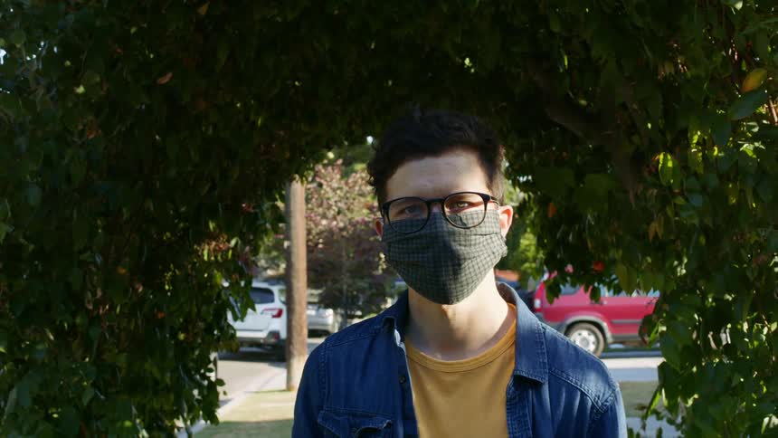 A man is standing on a neighborhood sidewalk staring into the camera while wearing a mask