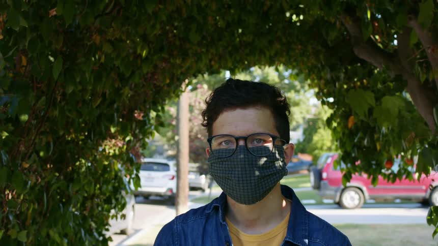 Young man wearing glasses and face mask stands under a trimmed bush in a parking lot