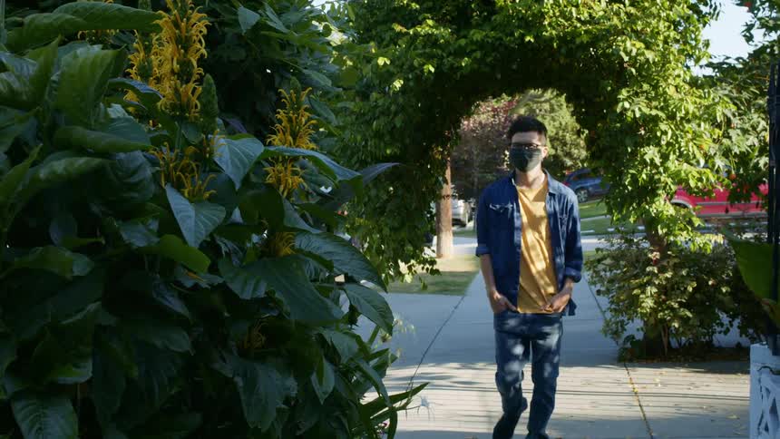 A man with glasses and a mask walking on a pavement with vehicles in the background