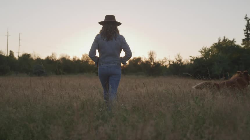 Woman with a cowboy hat a shirt and jeans walks a brown dog through tall dry grass in a field at sunset