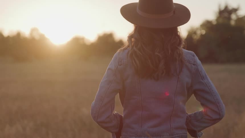 A woman with a hat and jeans jacket standing in a grassy field with trees and the sky in the background