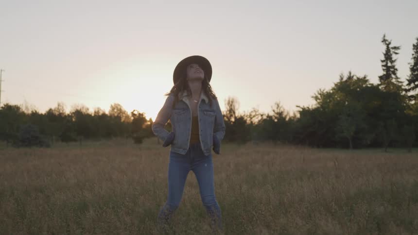 Woman stands in field turning and looking up as sunshine hovers over woods on horizon