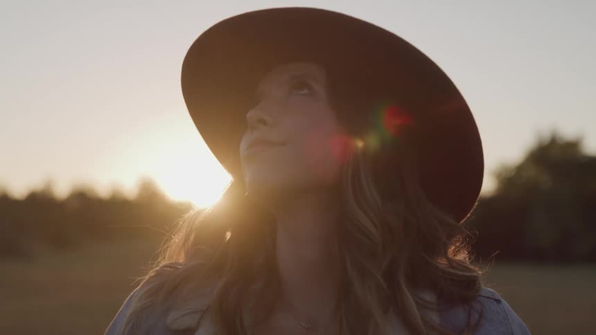 A teenage girl in a field looks up at the sky as the sun sets behind her