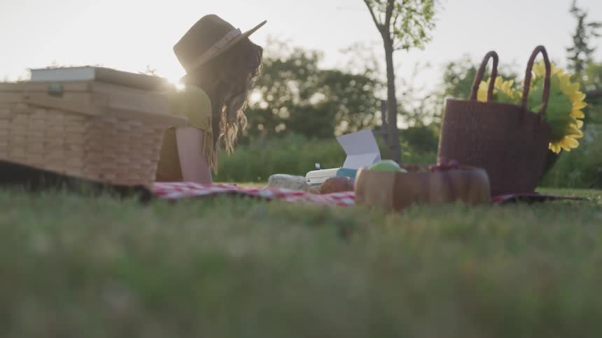 Woman wearing a hat lays over a blanket next to a picnic basket over the grass at sunset