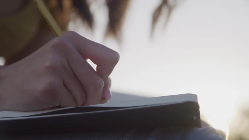 Womans hand writes in a notebook outdoors at sunset