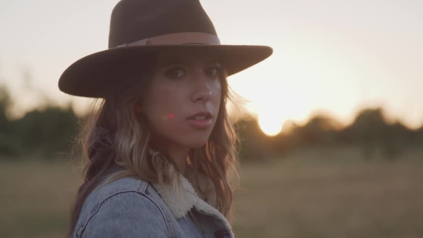 A young woman in a hat and jacket turns to look into the camera as the sun sets behind her