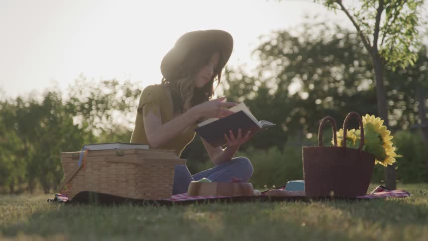 Young woman reading a book on picnic blanket with basket on lawn